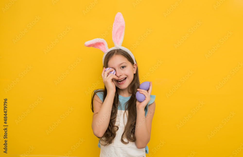 A cute girl with dark hair in a headband with rabbit ears holds a purple Easter egg in one hand in front of her eyes and smiles while looking at the camera.Studio portrait on a yellow background
