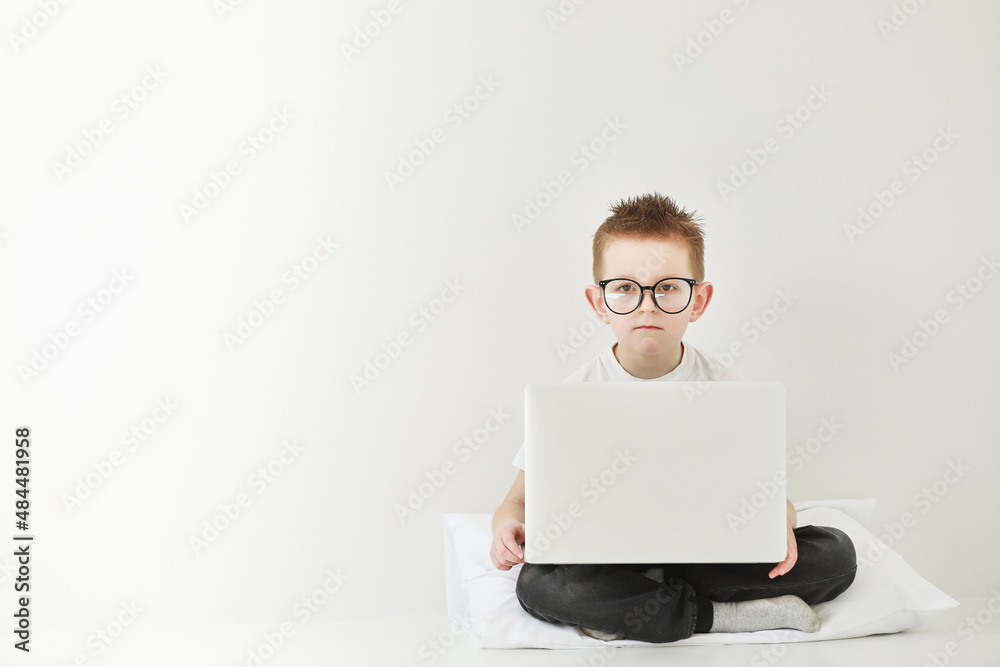 schoolboy in glasses using laptop sitting on bed. Child doing school homework and seriously and tired looking on camera.Online learning, distance lesson, education with online tutor at home isolation schoolboy in glasses using laptop sitting on bed. Child doing school homework and seriously and tired looking on camera.Online learning, distance lesson, education with online tutor at home isolation