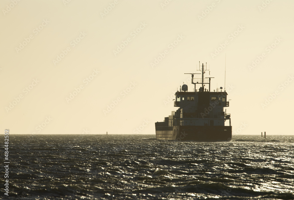 silhouette of big cargo ship sailing with the full power out to the ocean in th evening, no clouds and yellow color sky. water transportation concept