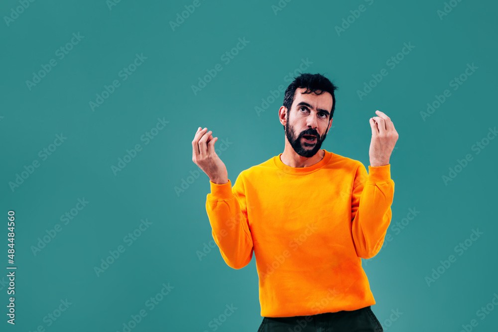 Italian man doing iconic Italian gesture of pine cone with hand - latin male with confused face questioning with hands - orange sweatshirt on green marine background Italian man doing iconic Italian gesture of pine cone with hand - latin male with confused face questioning with hands - orange sweatshirt on green marine background