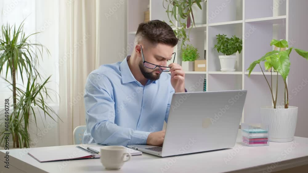 Tired of working at a laptop, a business man takes off his glasses while sitting at a work desk.