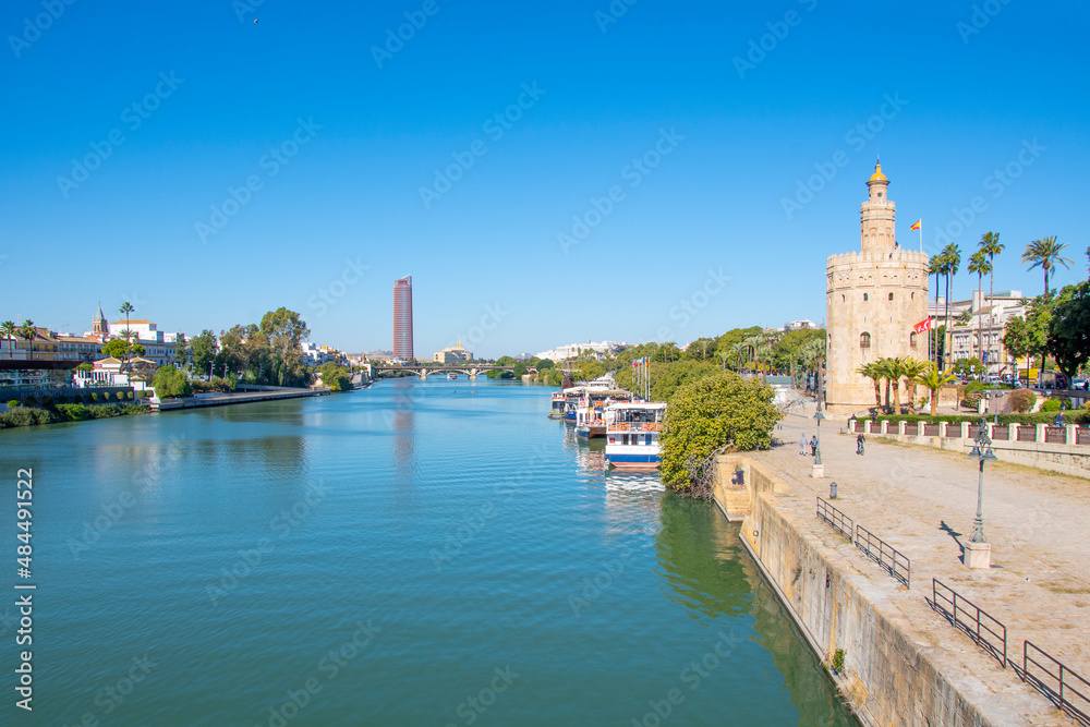 The Guadalquivier River as it runs through the historic center of Seville, Spain with the Triana district on one side, the Torre del Oro watchtower on the other, and the Sevilla Tower in the distance.