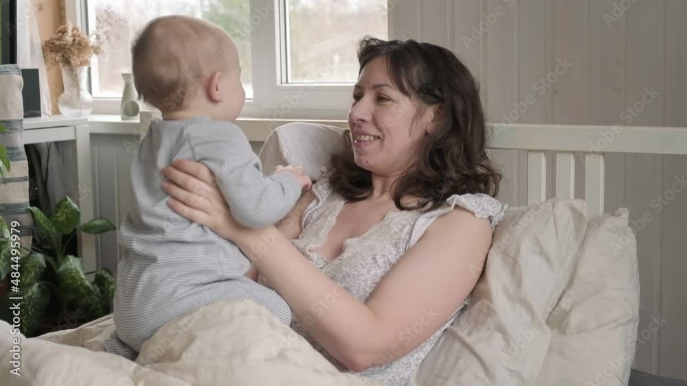 One Caucasian brunette mother in nightwear waking up with her tiny newborn, playing on bed. Young woman with baby relaxing at home. Bed-sharing concept. Infant child with his mom in bedroom. One Caucasian brunette mother in nightwear waking up with her tiny newborn, playing on bed. Young woman with baby relaxing at home. Bed-sharing concept. Infant child with his mom in bedroom.