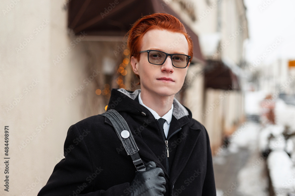 Stylish handsome young redhead hipster man with glasses in a black jacket with a backpack walks in the city. Business traveler in fashionable outerwear on a winter day. Male portrait