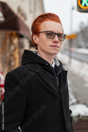 Urban portrait of a beautiful young red-haired man with glasses in fashionable business clothes with a coat, shirt and tie walks on the street.