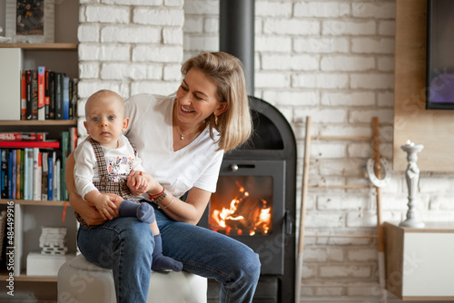 Fototapet Portrait of smiling blond woman with baby in hands basking near comfort hot warm fireplace with firewood burning inside