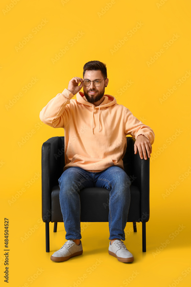 Handsome bearded man with eyeglasses sitting in leather armchair on yellow background Handsome bearded man with eyeglasses sitting in leather armchair on yellow background