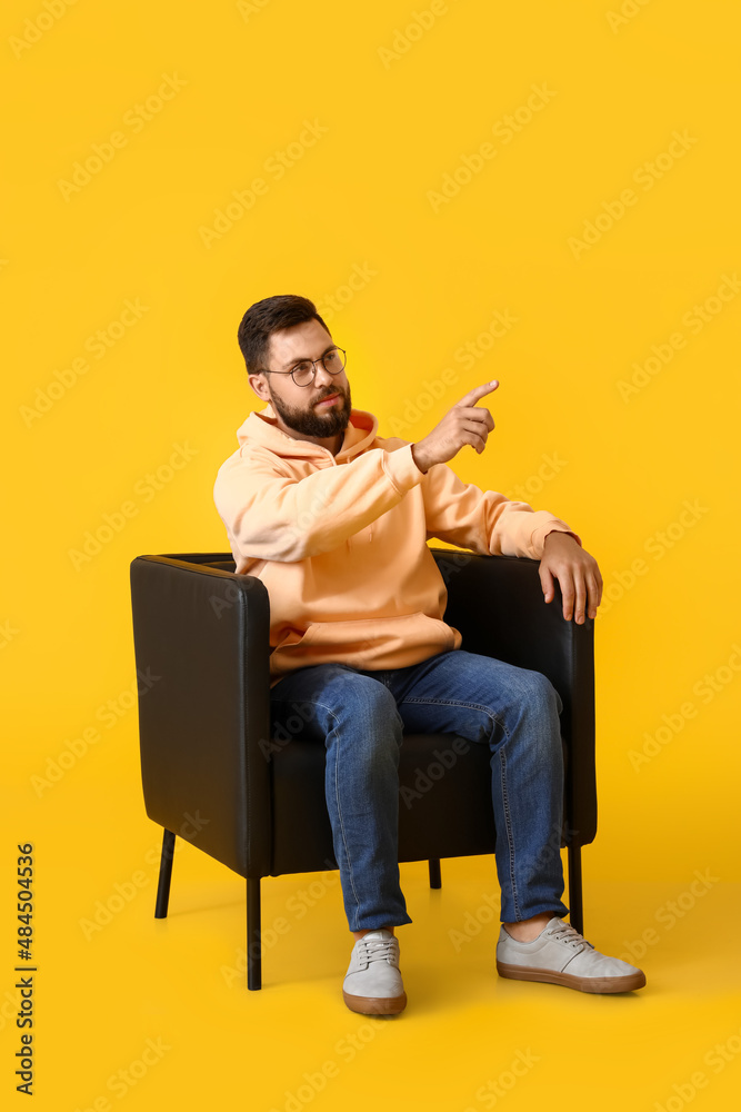 Handsome bearded man in leather armchair pointing at something on yellow background