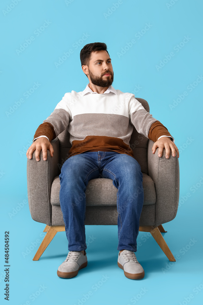 Handsome bearded man sitting in soft armchair on blue background