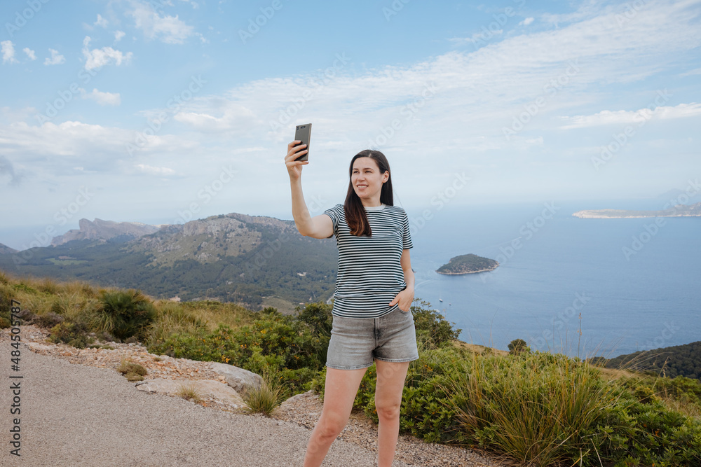 Woman in shorts takes a selfie on the mountain with panoramic view in Italy, Spain or Greece. Summer vacation by the sea. Take pictures on vacation. 