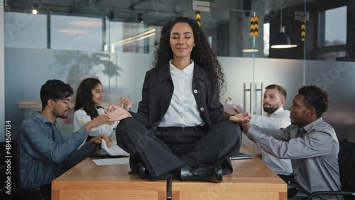 Young hispanic girl sitting on table in office in lotus position relaxing feeling calm, harmony businesswoman meditate ignore angry annoying colleagues female executive doing yoga exercises for stress photo