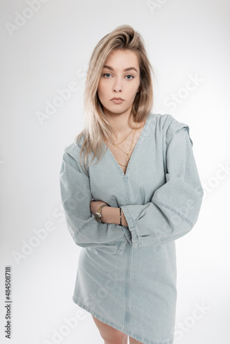 Stylish beautiful blonde woman with hairstyle in trendy denim clothes stands in the studio. Fashion girl