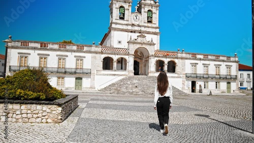 Beautiful young girl walking by a Catholic Church in Nazaré Miradouro do Suberco. A tourist woman walking in slow motion and exploring art and architecture in an old Town in Portugal, Europe.