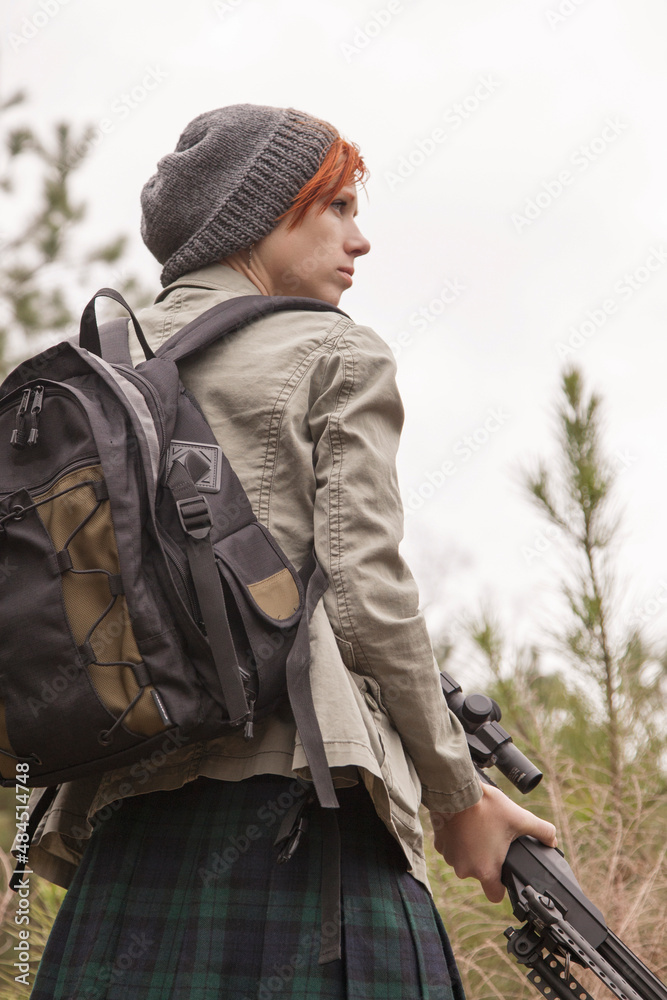 Fototapeta premium Beautiful and intense redhead sniper woman in profile with .50 caliber rifle pointed down, short hair, stocking cap, jacket, backpack looks sideways through the forest on an overcast day, low angle