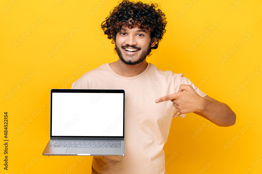 Positive confident Indian stylish curly guy, holds an open laptop with blank white screen, points finger at it, standing on isolated orange background, smiling friendly. Copy space, mock-up concept Positive confident Indian stylish curly guy, holds an open laptop with blank white screen, points finger at it, standing on isolated orange background, smiling friendly. Copy space, mock-up concept