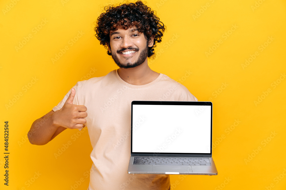 Excited cheerful Indian or Arabian guy in basic t-shirt, holds open laptop with blank white screen, showing thumbs up gesture, standing on isolated orange background, friendly smiling. Mock-up concept Excited cheerful Indian or Arabian guy in basic t-shirt, holds open laptop with blank white screen, showing thumbs up gesture, standing on isolated orange background, friendly smiling. Mock-up concept