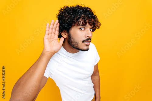 Deafness  gossip concept. Positive Indian man  wearing basic t-shirt with hand near ear concentrated listening rumor  hearing gossip  standing on isolated orange background
