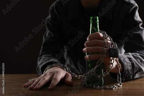 Alcohol addiction. Man chained with bottle of beer at wooden table, closeup