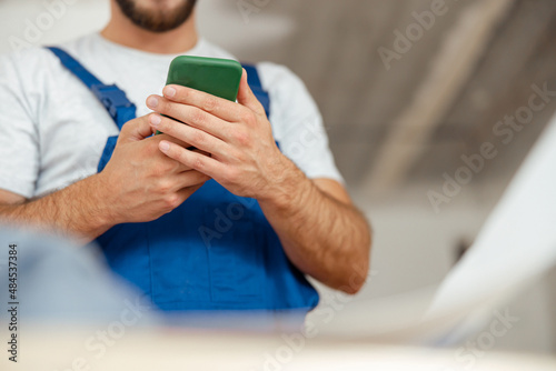 Closeup of hands of repairman in workwear texting, using smartphone, standing indoors during renovation work