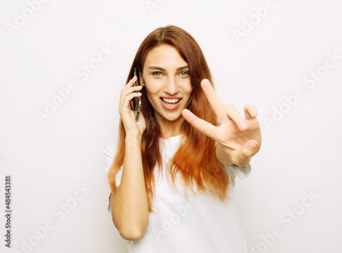 Young redhead woman holding mobile phone and carefree showing a peace symbol with fingers.
