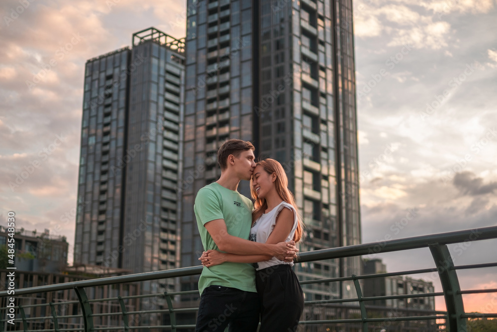 Lovely young couple standing on the street in the metropolis on the sunset and cuddling. Boyfriend kisses girlfriend. Skyscrapers on the background