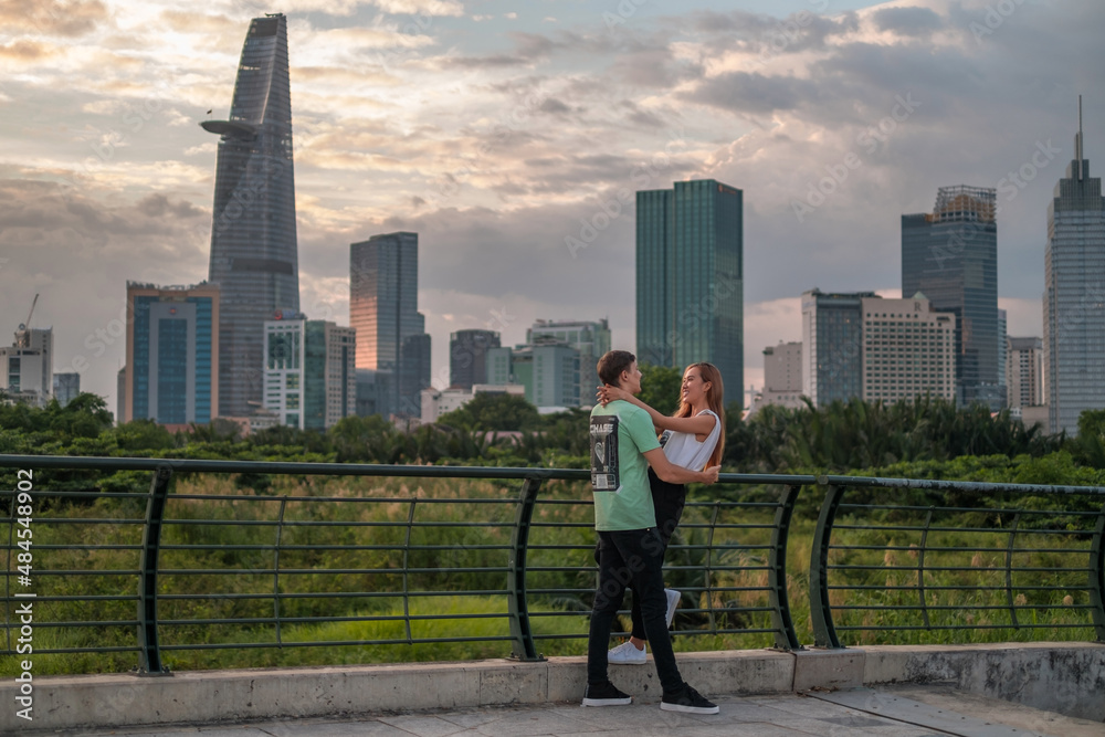 Lovely mixed couple standing on the bridge in the park in metropolis and cuddling. Love people and happiness concept. Skyscrapers on the background Lovely mixed couple standing on the bridge in the park in metropolis and cuddling. Love people and happiness concept. Skyscrapers on the background