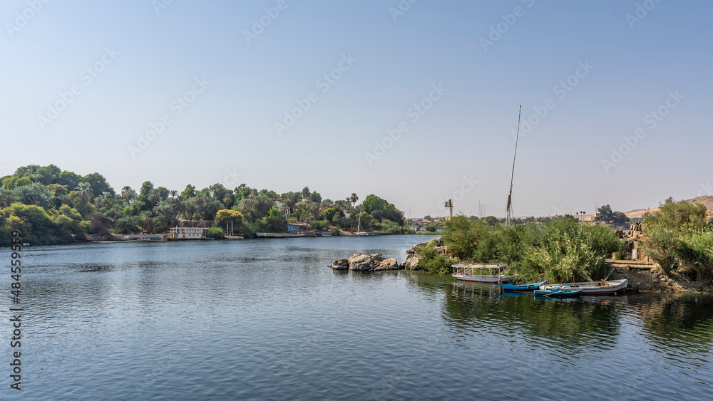 On the banks of the Nile, lush green vegetation, picturesque boulders. The boats are on calm water. Clear blue sky. Reflection. Egypt