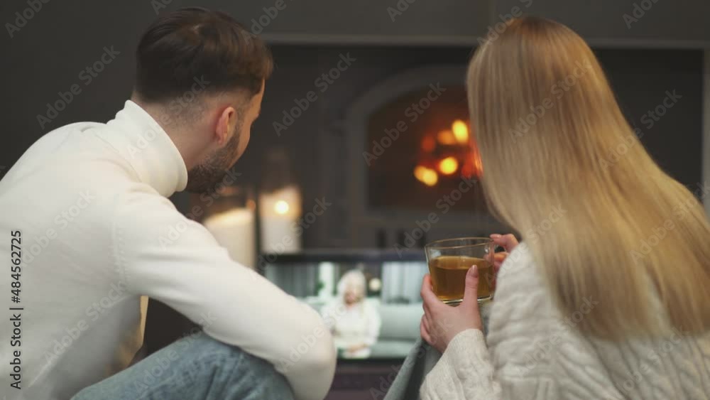 Handheld, young couple sits by the fireplace in living room and communicates with grandmother on a video call using a laptop, communication at a distance. Handheld, young couple sits by the fireplace in living room and communicates with grandmother on a video call using a laptop, communication at a distance.