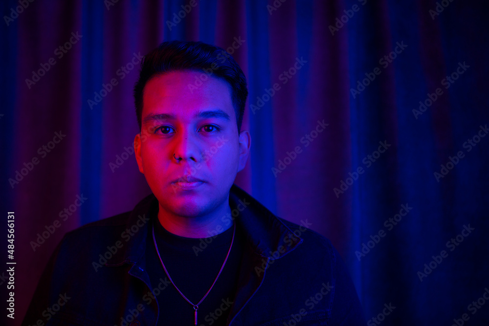 Young latin man headshot under blue and red lights. He is serious and looking to the camera. Young latin man headshot under blue and red lights. He is serious and looking to the camera.