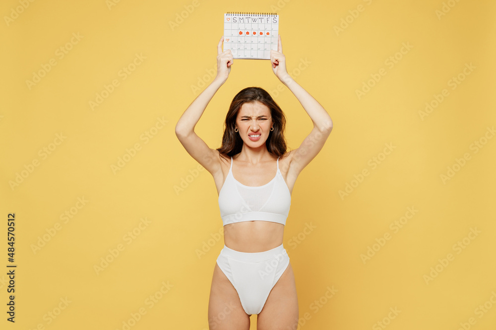 Naklejka premium Sad dissatisfied young woman 20s in white underwear with fit body hold in hand female periods calendar checking menstruation days isolated on plain yellow background Healthcare, gynecological concept