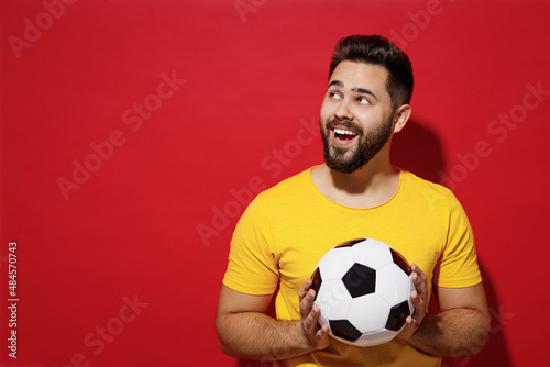 Fun young bearded man football fan in yellow t-shirt support favorite team look up on workspace area copy space mock up hold in hands soccer ball isolated on plain dark red background studio portrait. © ViDi Studio