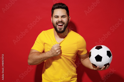 Fancy young bearded man football fan pull tear yellow t-shirt cheer up support favorite team hold soccer ball isolated on plain dark red background studio portrait. People emotions lifestyle concept.
