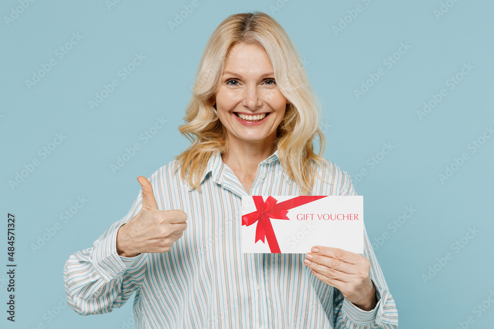 Elderly smiling fun woman 50s in striped shirt hold gift certificate coupon voucher card for store show thumb up isolated on plain pastel light blue color background studio. People lifestyle concept. Elderly smiling fun woman 50s in striped shirt hold gift certificate coupon voucher card for store show thumb up isolated on plain pastel light blue color background studio. People lifestyle concept.