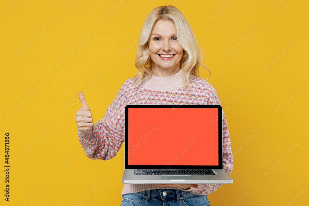 Elderly happy smiling blonde woman 50s in pink sweater hold use work on laptop pc computer with blank screen workspace area show thumb up gesture isolated on plain yellow background studio portrait Elderly happy smiling blonde woman 50s in pink sweater hold use work on laptop pc computer with blank screen workspace area show thumb up gesture isolated on plain yellow background studio portrait