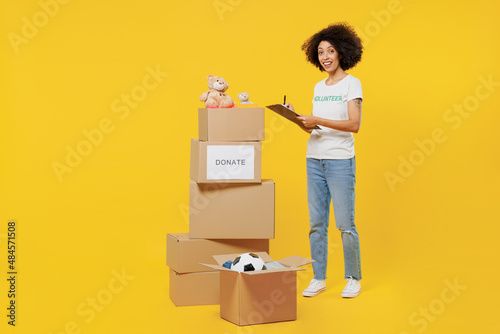 Full size young woman of African American ethnicity in white volunteer t-shirt hold boxes with presents writing list isolated on plain yellow background. Voluntary free work assistance help concept. photo