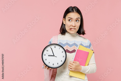 Confused sad unhappy teen student girl of Asian ethnicity wearing sweater backpack hold books clock isolated on pastel plain light pink background Education in high school university college concept.