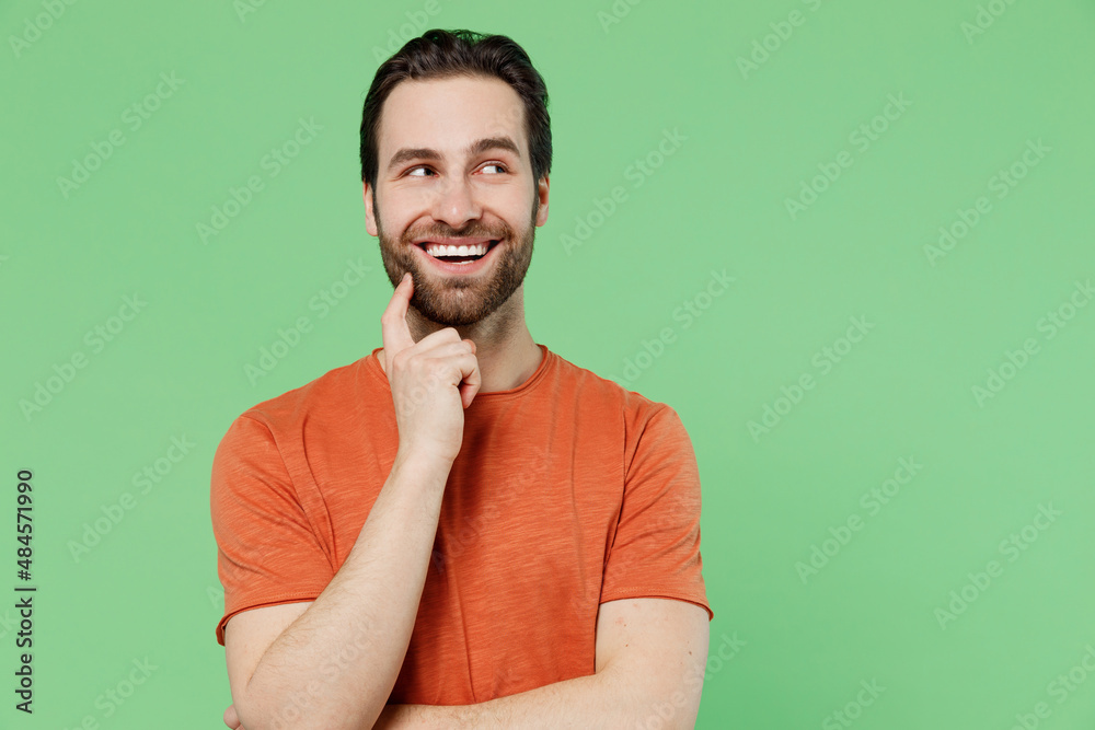 Young minded man 20s wearing casual orange t-shirt prop up chin look aside on workspace area mock up copy space isolated on plain pastel light green color background studio. People lifestyle concept. Young minded man 20s wearing casual orange t-shirt prop up chin look aside on workspace area mock up copy space isolated on plain pastel light green color background studio. People lifestyle concept.