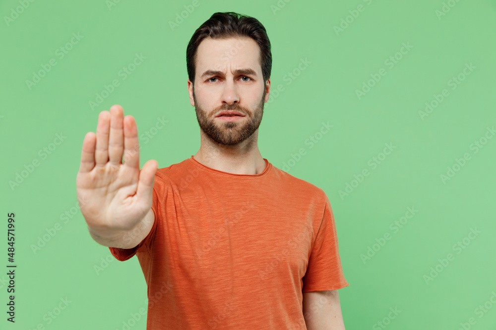 Young strict sad caucasian man 20s wearing casual orange t-shirt showing stop gesture with palm refuse isolated on plain pastel light green color background studio portrait. People lifestyle concept. Young strict sad caucasian man 20s wearing casual orange t-shirt showing stop gesture with palm refuse isolated on plain pastel light green color background studio portrait. People lifestyle concept.