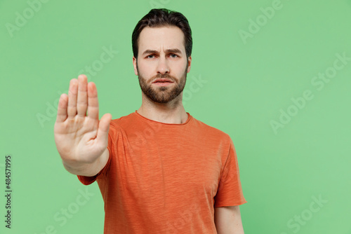 Young strict sad caucasian man 20s wearing casual orange t-shirt showing stop gesture with palm refuse isolated on plain pastel light green color background studio portrait. People lifestyle concept.