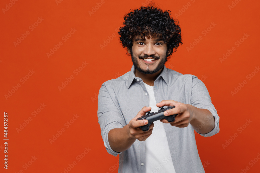 Gambling overjoyed cheerful excited jubilant young bearded Indian man 20s years old wear blue shirt hold in hand play pc game with joystick console isolated on plain orange background studio portrait Gambling overjoyed cheerful excited jubilant young bearded Indian man 20s years old wear blue shirt hold in hand play pc game with joystick console isolated on plain orange background studio portrait