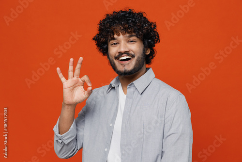 Cheerful exultant happy overjoyed young bearded Indian man 20s years old wear blue shirt showing okay ok gesture isolated on plain orange background studio portrait. People emotions lifestyle concept © ViDi Studio