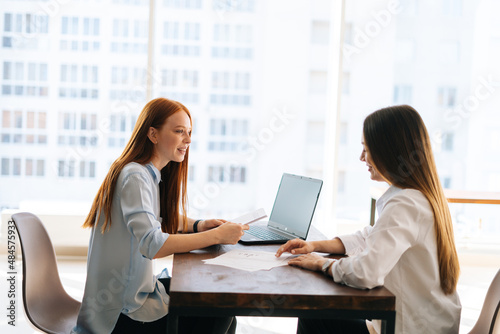 Side view of two young female colleagues discuss financial data in report using laptop and paper documents, sitting at desk opposite each other in office, on background of window and city buildings.