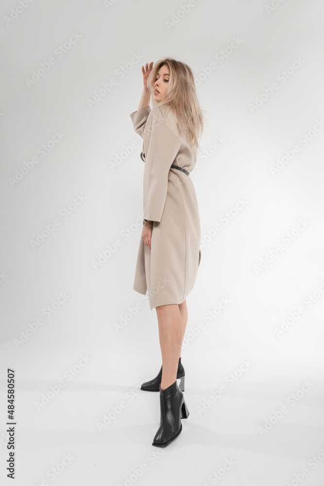 Pretty sexy young woman model with fashion classic beige coat in black leather shoes poses in studio on a white background. Stylish new spring collection of outwear Pretty sexy young woman model with fashion classic beige coat in black leather shoes poses in studio on a white background. Stylish new spring collection of outwear
