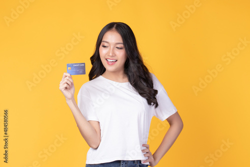 Portrait of a happy young asian woman wearing white shirt holding bank card, credit card isolated on yellow background. Business online shopping concept.