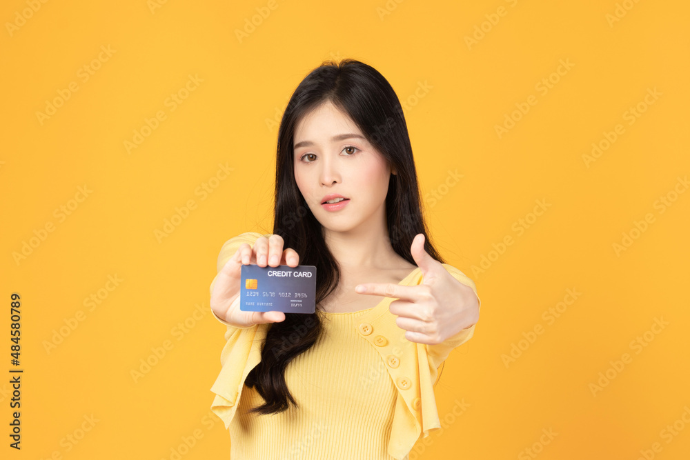 Cheerful young asian woman in yellow T-shirt holding bank card isolated on yellow background. Asian girl point to credit card. Cheerful young asian woman in yellow T-shirt holding bank card isolated on yellow background. Asian girl point to credit card.