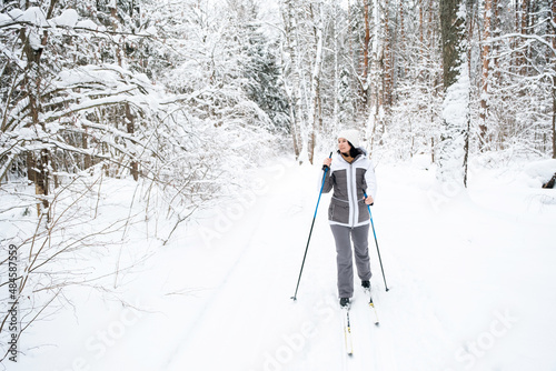 Skier a woman in a membrane jacket with ski poles in his hands with his back against the background of a snowy forest. Cross-country skiing in winter forest, outdoor sports, healthy lifestyle.
