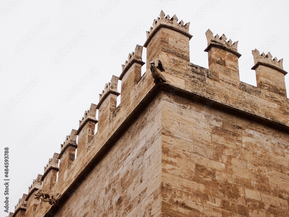 The wall of an old stone building with battlements on the roof and a statue of a gargoyle in the form of a winged monster. Medieval towers and castles, historical and ancient cities. Soft focus The wall of an old stone building with battlements on the roof and a statue of a gargoyle in the form of a winged monster. Medieval towers and castles, historical and ancient cities. Soft focus