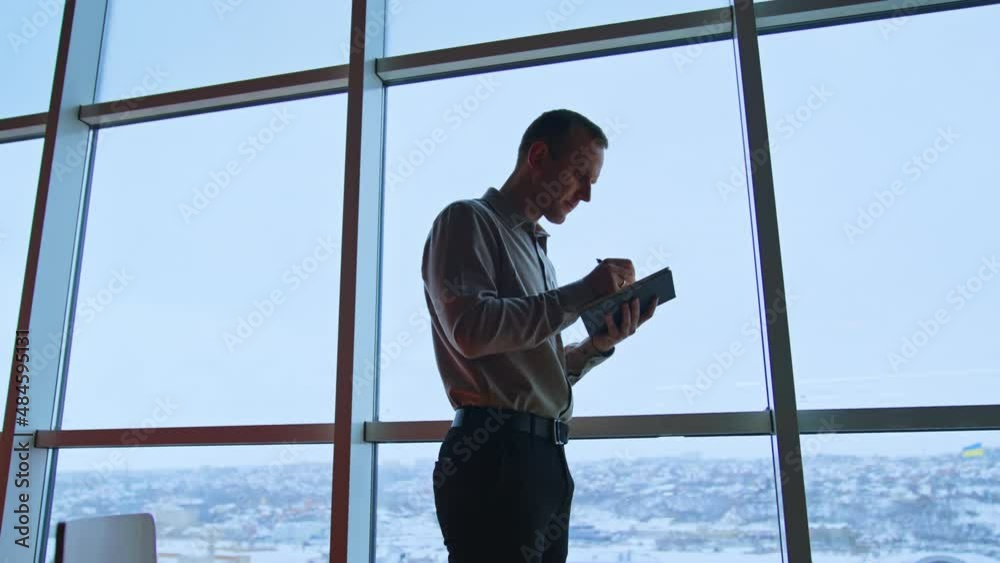 Tall slim man stands against the panoramic window. Male taking notes, looks into window and throws away one page. City view at backdrop.