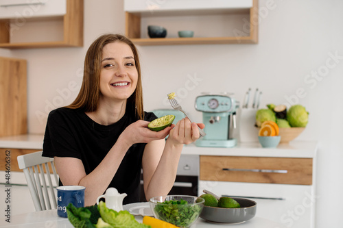Young beautiful vegetarian girl dressed in pajamas eating fruits and vegetables for breakfast at home in the kitchen