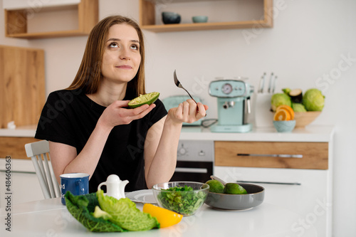 Young beautiful vegetarian girl dressed in pajamas eating fruits and vegetables for breakfast at home in the kitchen